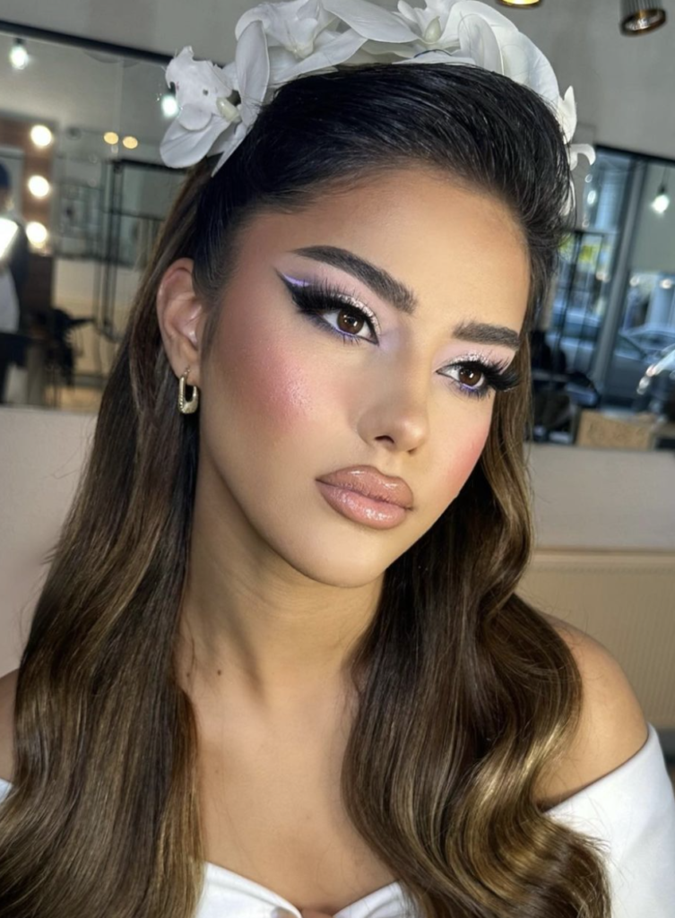A woman with a bridal makeup look, featuring a sharp winged eyeliner in a soft lilac shade, framed by white floral hair accessories. Her cheeks are adorned with a rosy blush, and her lips are glossy with a pale pink hue. Her long brunette hair flows down in soft waves, complementing her ethereal appearance.